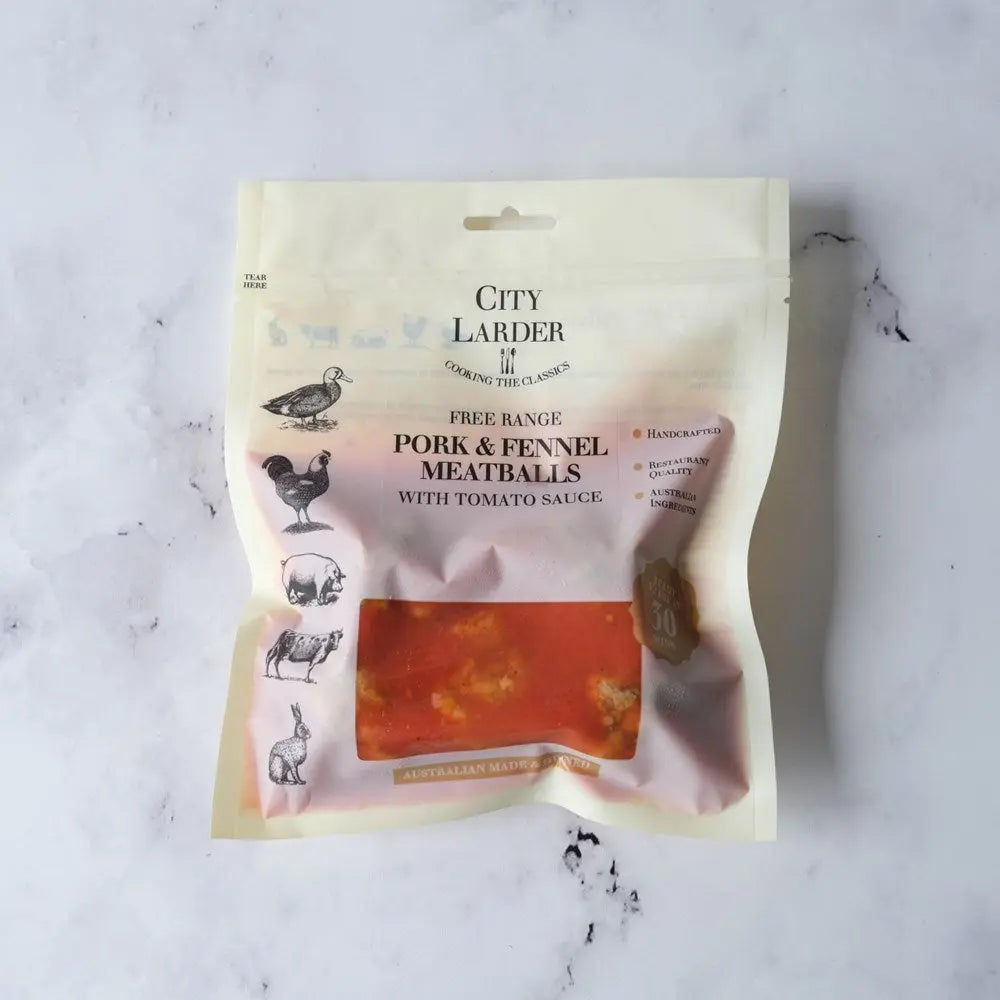 A stand-up pouch of City Larder Free Range Pork & Fennel Meatballs with Tomato Sauce, displayed on a light gray marble surface. The packaging is cream-colored with black text and illustrations of various farm animals, and features a clear window showing the orange-red meatballs inside.