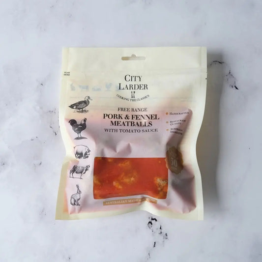 A stand-up pouch of City Larder Free Range Pork & Fennel Meatballs with Tomato Sauce, displayed on a light gray marble surface. The packaging is cream-colored with black text and illustrations of various farm animals, and features a clear window showing the orange-red meatballs inside.